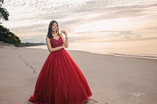 Quinceañera en vestido rojo durante una sesión de fotos en la playa al amanecer, hora dorada.