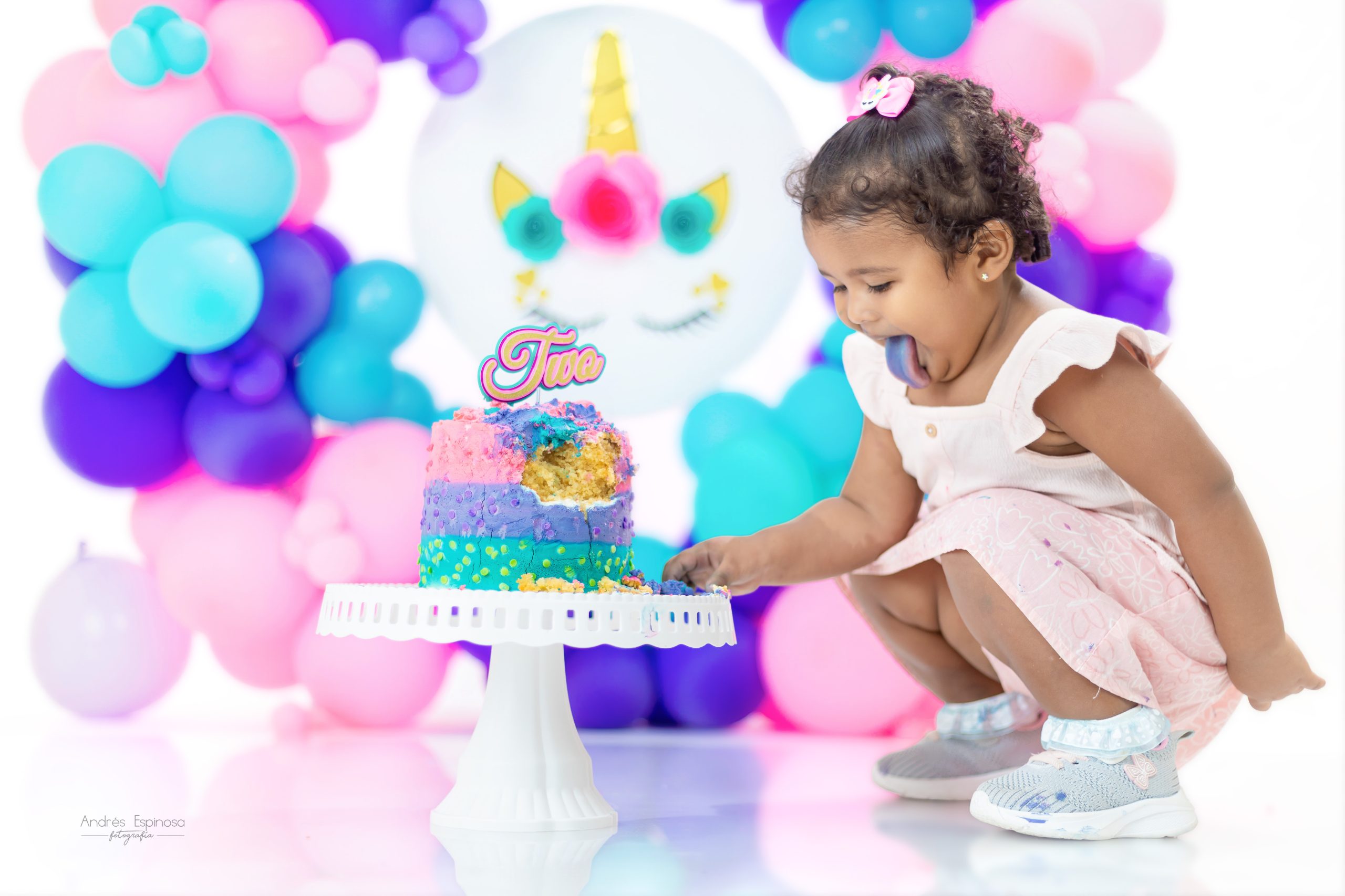 Niña pequeña en sesión smash cake probando su tarta de colores frente a un fondo de globos rosas, azules y morados.