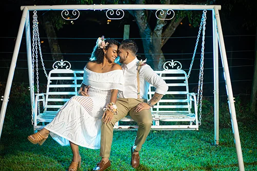Pareja sonriendo en un columpio de hierro blanco en un jardín nocturno, ella con vestido blanco y corsage floral, él con tirantes y camisa blanca.