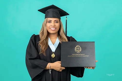 Graduada sonriente en toga y birrete sosteniendo su diploma de la Universidad de Panamá sobre fondo turquesa.