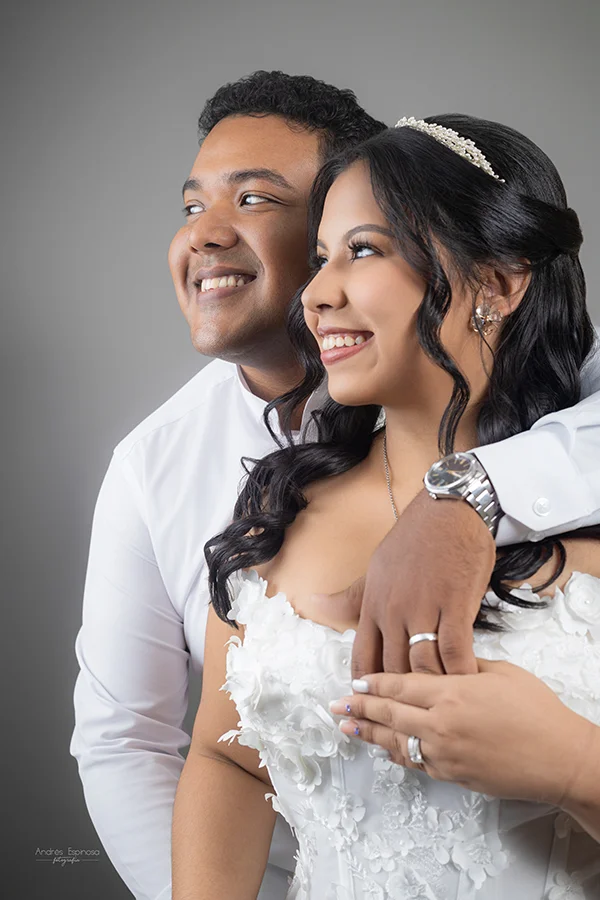 Pareja en sesión preboda: él abrazándola por detrás mientras sonríen, ella luce un vestido blanco con aplicaciones florales y tiara, sobre fondo neutro.