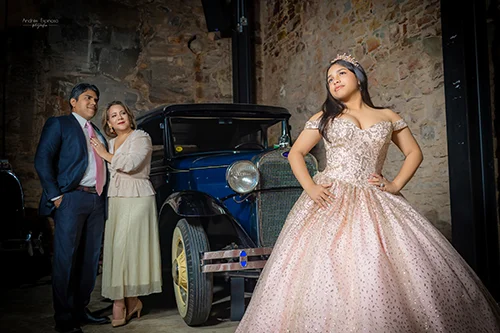 Quinceañera en vestido rosa brillante posando con las manos en cintura junto a un automóvil clásico, con sus padres admirándola al fondo.